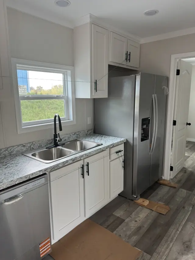 Kitchen corner with a double sink under a window, white cabinets, and a stainless steel side-by-side refrigerator beside a hallway door.