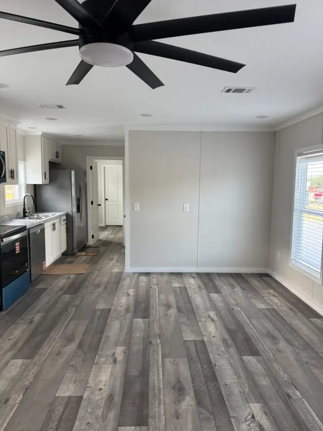 Open living area with a black ceiling fan, gray plank flooring, and the kitchen and a hallway in the background.